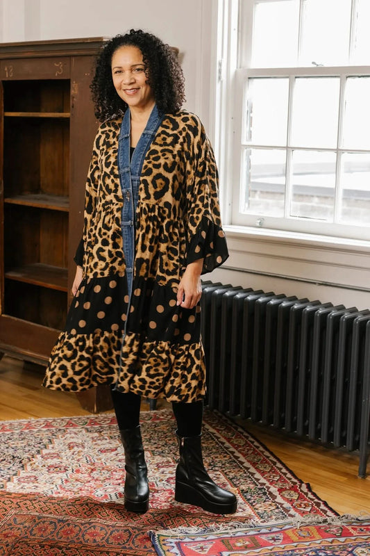 A woman stands against a stone wall wearing a black shirt, upcycled denim jeans, black boots, and the Shannon Passero Ying Cardigan—an eye-catching long coat with animal print and polka dots.