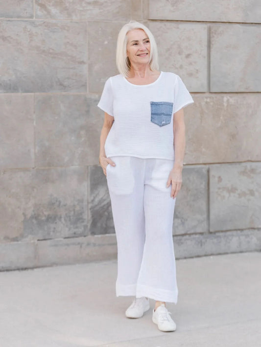 An older woman with straight white hair wears the Aletta Top by Shannon Passero, a cotton gauze shirt with a blue upcycled vintage denim pocket, paired with white pants and standing against a stone wall.