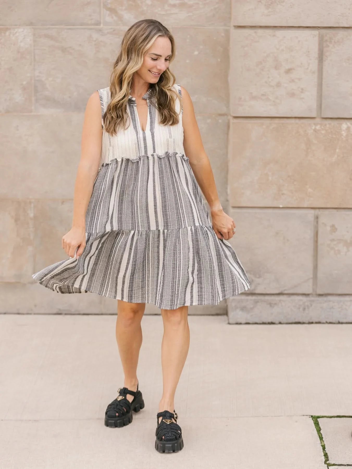 A person with long hair stands outdoors in a playful, v-neck, sleeveless Shannon Passero Whitby Tank Dress, smiling slightly.