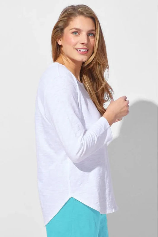 A woman wearing the Escape Peruvian Cotton Weekender Tee in white and light blue pants stands against a plain light background, looking down as she touches her hair.
