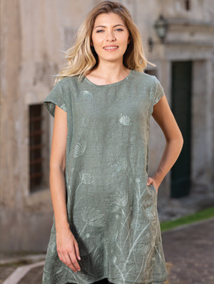 A woman stands outdoors wearing the Cherishh Short Dress With Emb, a short-sleeve green tunic featuring a subtle botanical pattern, with one hand in her pocket.