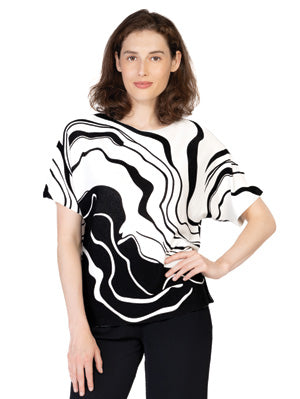 A woman stands facing forward, wearing the Variations Flair Top—a black and white short-sleeve top with an abstract wavy pattern—paired with black pants.