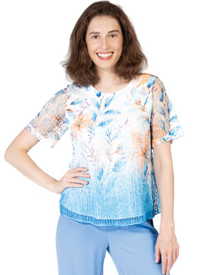 A woman wears the Variations Lace Top, a short-sleeve blue and white floral blouse, paired with light blue pants, standing and smiling at the camera with one hand on her hip.