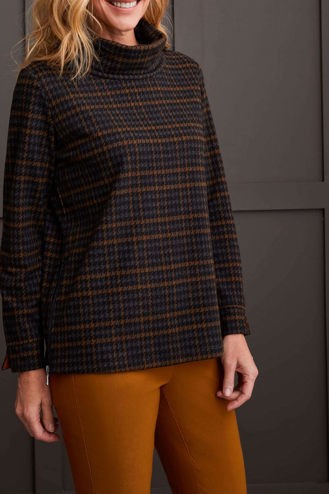 The person is wearing a chic cowl neck top with a side zipper by Tribal, paired with brown pants, standing comfortably in front of a gray paneled wall.
