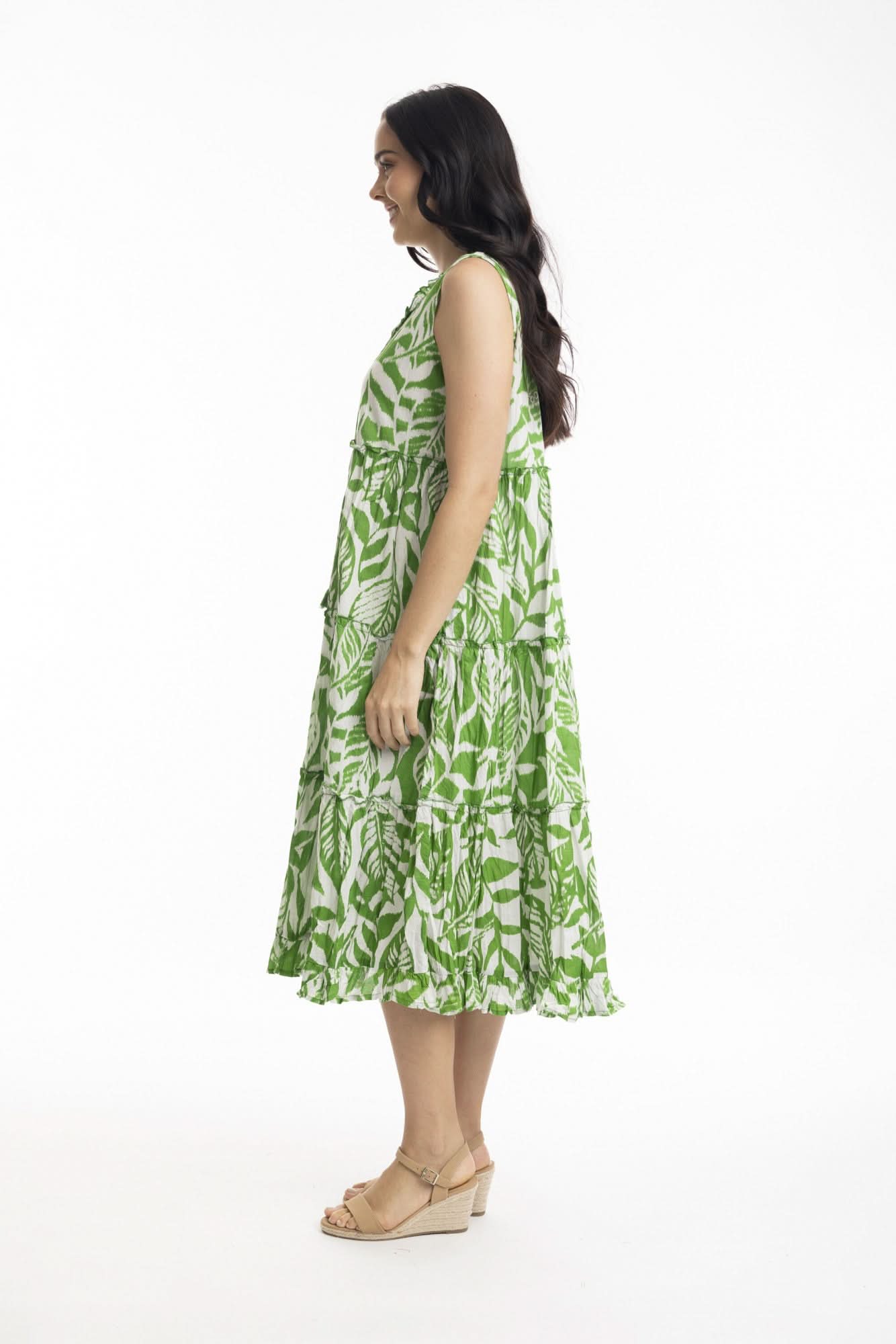 A woman smiles in the Orientique Hayman Green Sleeveless Boho Dress, featuring a green and white leaf pattern and tiered skirt, styled with beige heeled sandals—ideal for summer days against a plain white background.