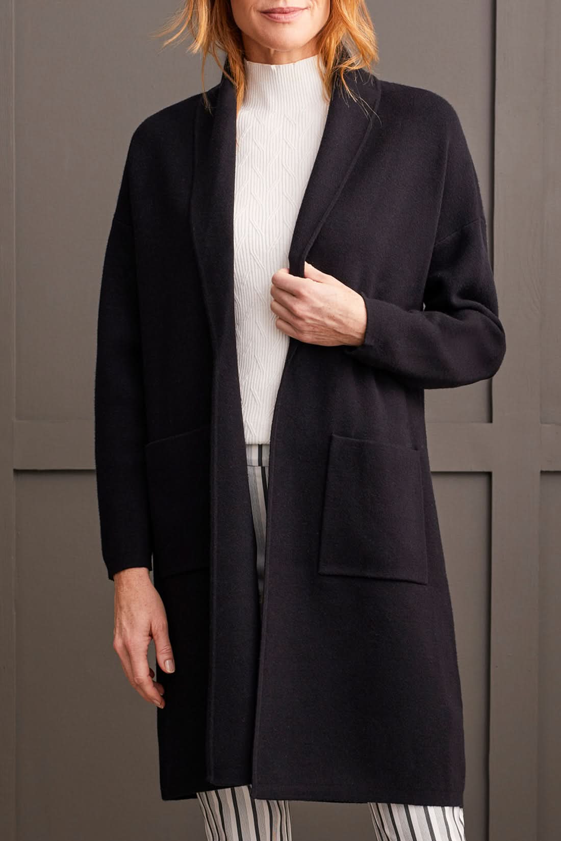 Individual wearing a stylish black Coatigan with Pockets by Tribal over a white high-neck sweater and striped pants, standing in front of a gray paneled wall.