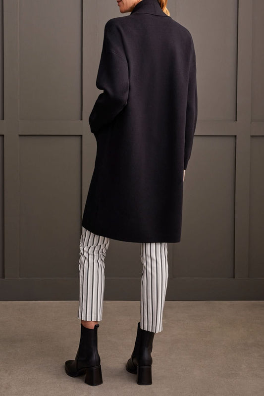 Individual wearing a stylish black Coatigan with Pockets by Tribal over a white high-neck sweater and striped pants, standing in front of a gray paneled wall.