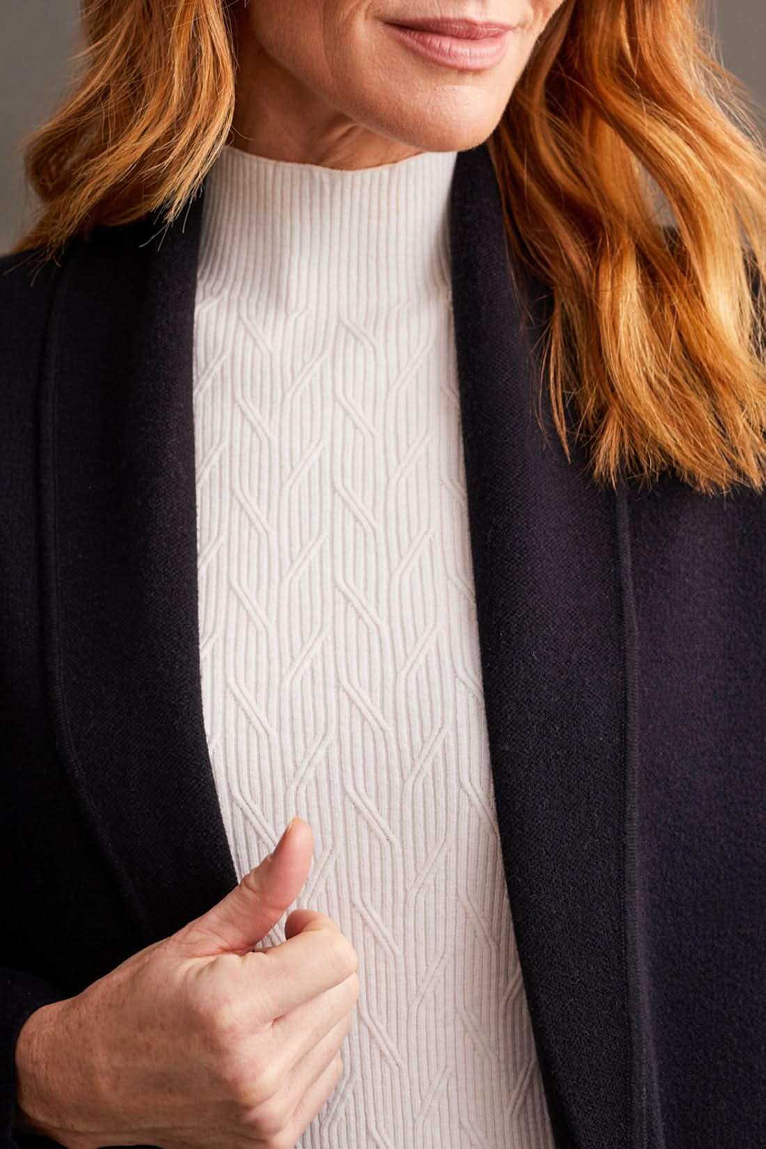 Individual wearing a stylish black Coatigan with Pockets by Tribal over a white high-neck sweater and striped pants, standing in front of a gray paneled wall.