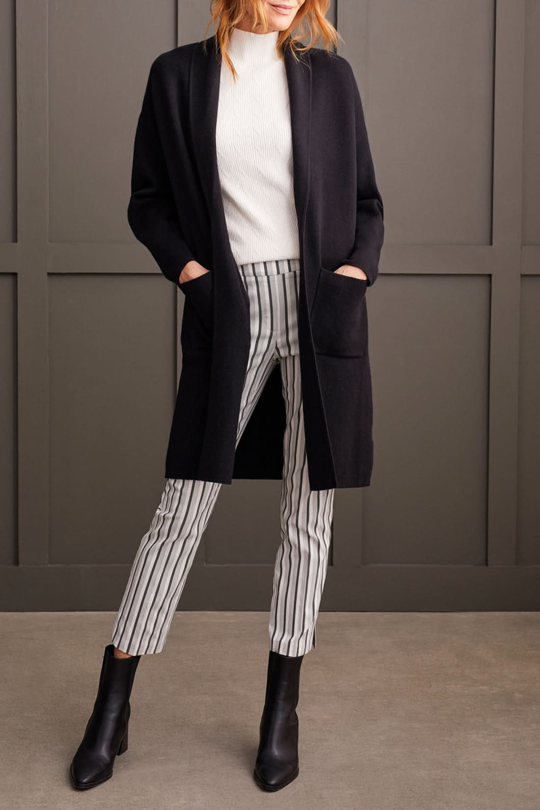 Individual wearing a stylish black Coatigan with Pockets by Tribal over a white high-neck sweater and striped pants, standing in front of a gray paneled wall.