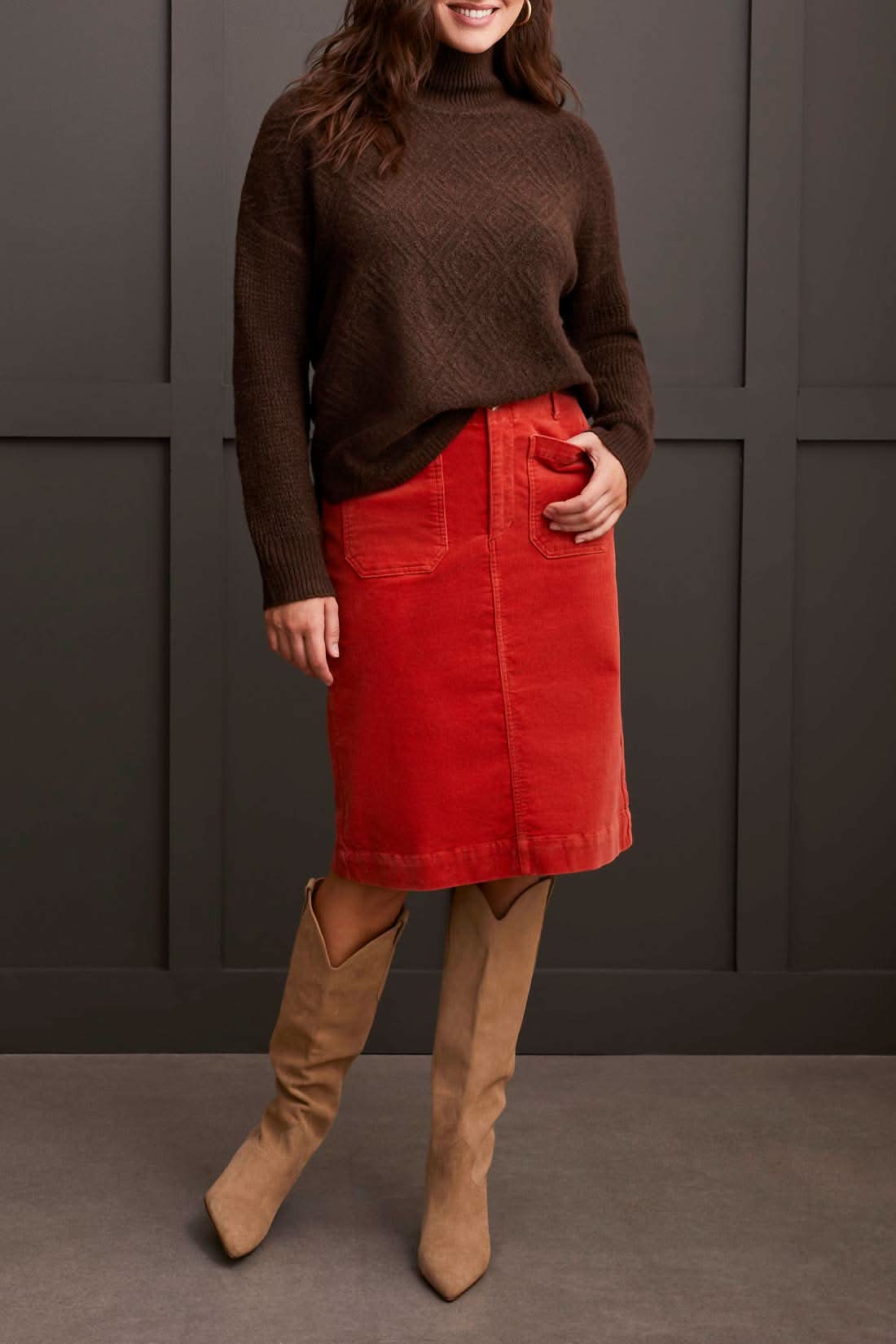 A person dressed in a cozy comfort ensemble featuring Tribal's coffee-colored Mock Neck Sweater, a red skirt, and tan knee-high boots stands against a dark paneled wall.