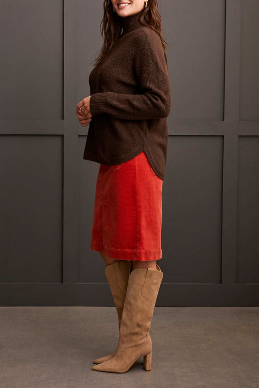 A person dressed in a cozy comfort ensemble featuring Tribal's coffee-colored Mock Neck Sweater, a red skirt, and tan knee-high boots stands against a dark paneled wall.