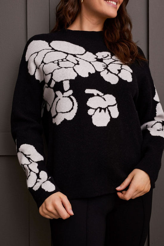 An individual is pictured from the shoulders down, donning a black Floral Funnel Neck Sweater by Tribal, adorned with a striking large white floral pattern. This cute fall sweater, made from cozy material, is ideal for cool autumn days. The person stands against a dark backdrop.