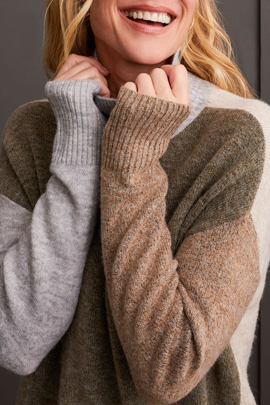 A person wearing the Mock Neck Colorblock Sweater by Tribal, featuring cozy gray, brown, and beige stripes, smiles while holding the collar near their face—truly a fall staple.