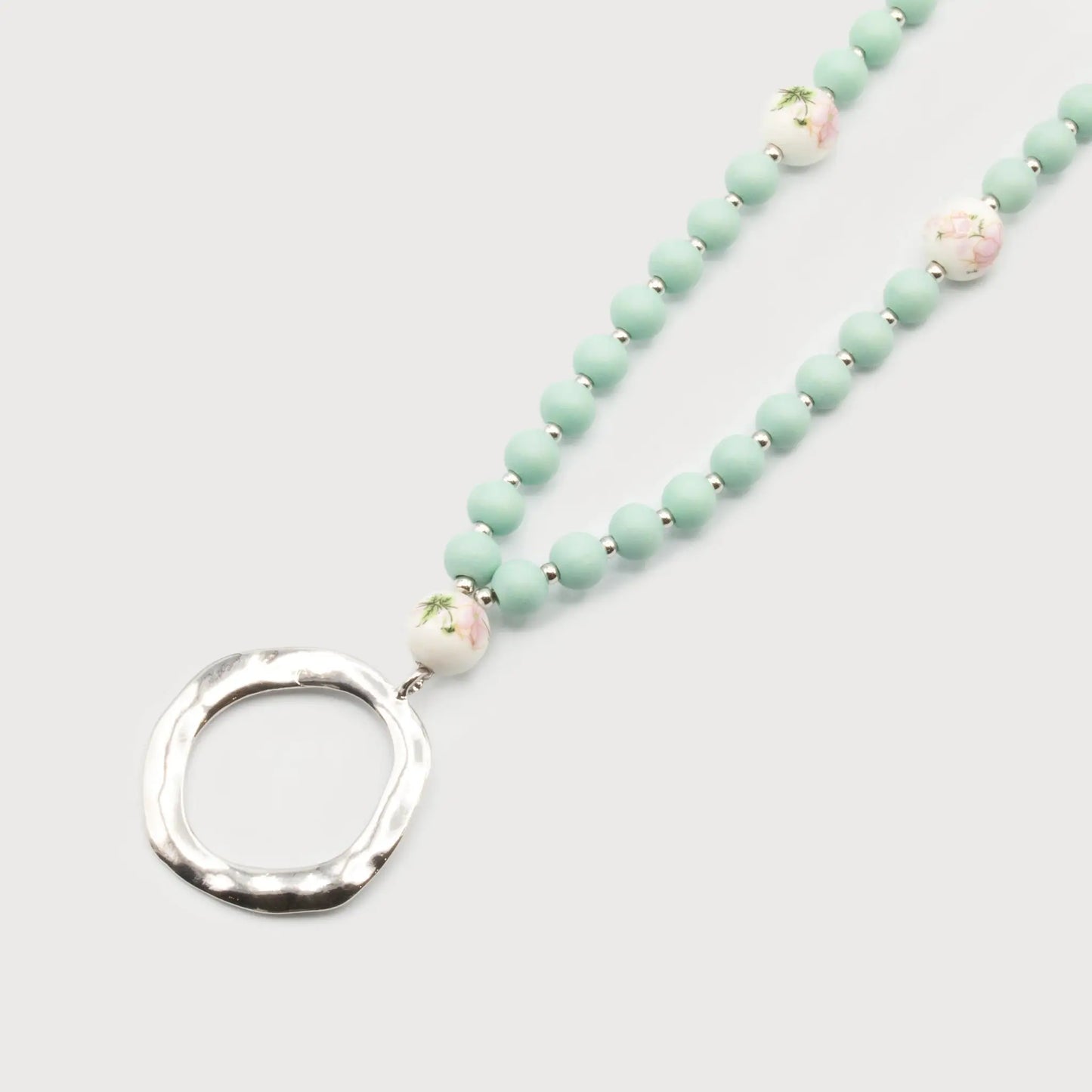 The Long Wood & Ceramic Beads Necklace by Caracol features mint green beads and decorative white ceramic beads with pink designs, all beautifully attached to a textured silver circular pendant.