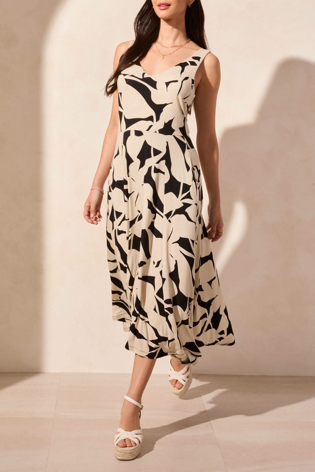 A woman stands against a light background, wearing Tribal's sleeveless high low dress featuring an abstract black and white print, paired with wedge sandals.