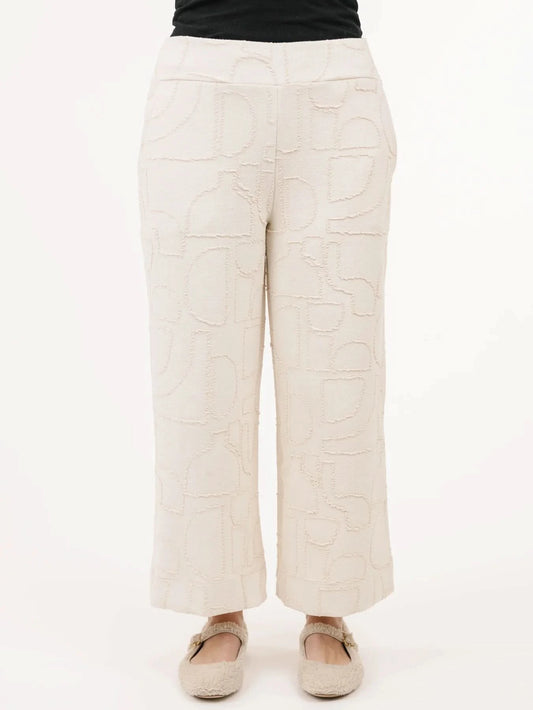 A woman stands indoors by a window on hardwood floors next to a radiator, wearing the Shannon Passero Newcastle Pant with a cream jacquard sweater and slippers.