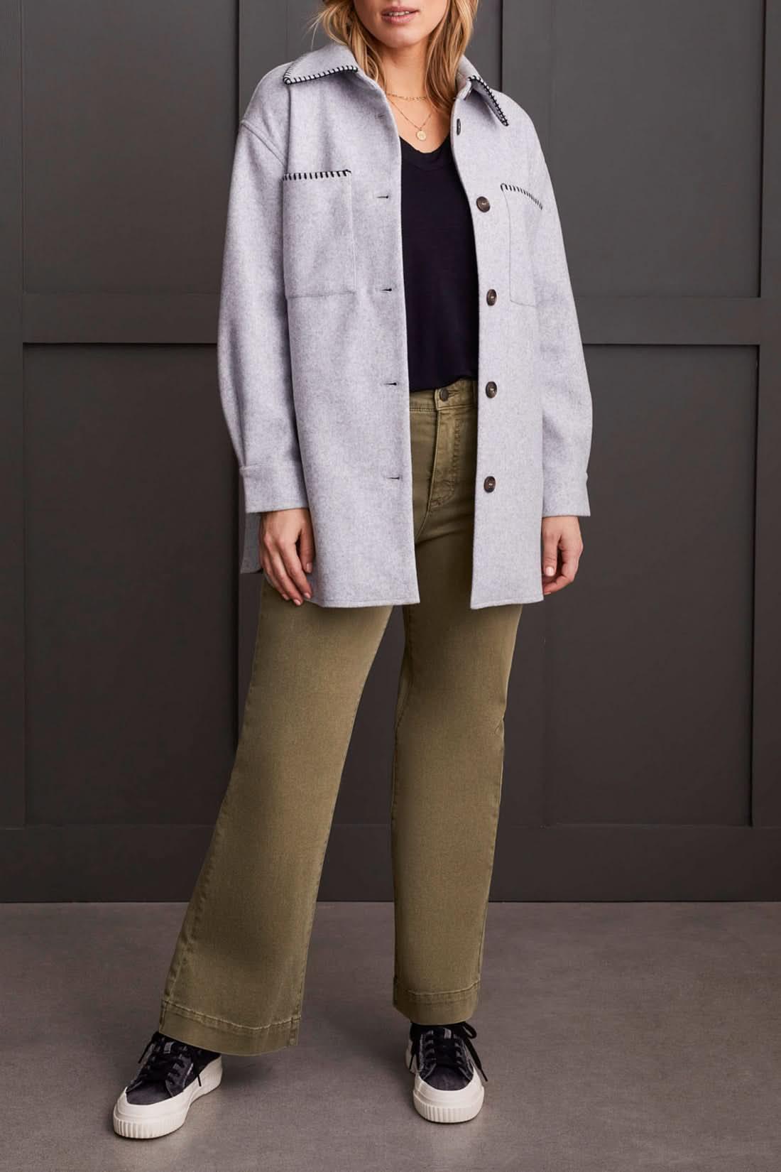 A person stands wearing the Tribal Button Front Coat with Blanket Stitch in light gray over a black top, green pants, and black-and-white sneakers, against a dark paneled background.