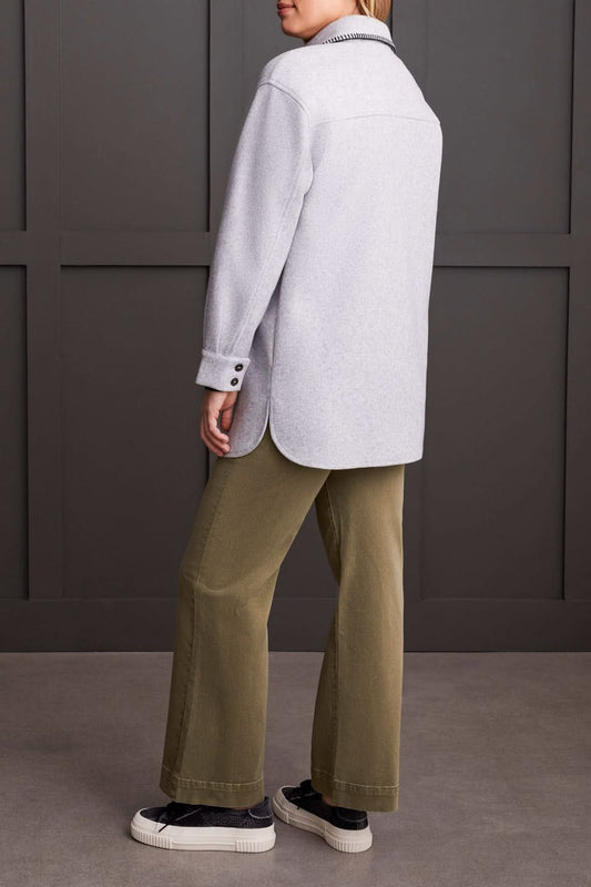 A person stands wearing the Tribal Button Front Coat with Blanket Stitch in light gray over a black top, green pants, and black-and-white sneakers, against a dark paneled background.