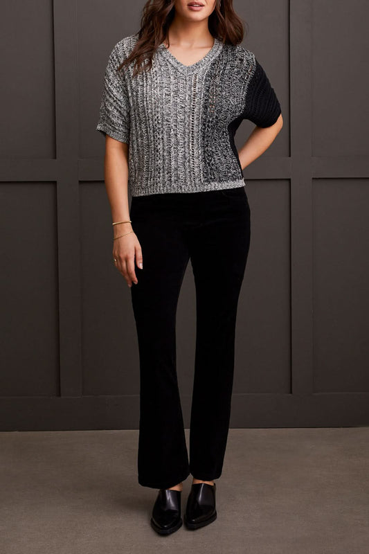A person wearing the Tribal Dolman Elbow Sleeve Sweater, a cozy dual-toned knit featuring black and gray sections, short Dolman elbow sleeves, and a V-neckline, is paired with black pants and stands against a dark background.
