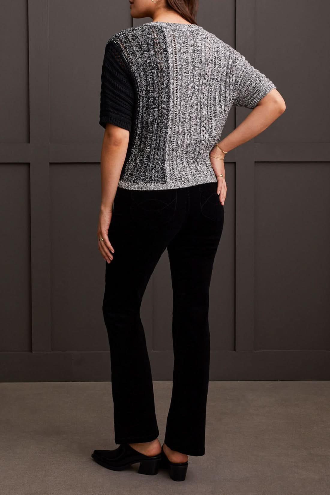 A person wearing the Tribal Dolman Elbow Sleeve Sweater, a cozy dual-toned knit featuring black and gray sections, short Dolman elbow sleeves, and a V-neckline, is paired with black pants and stands against a dark background.