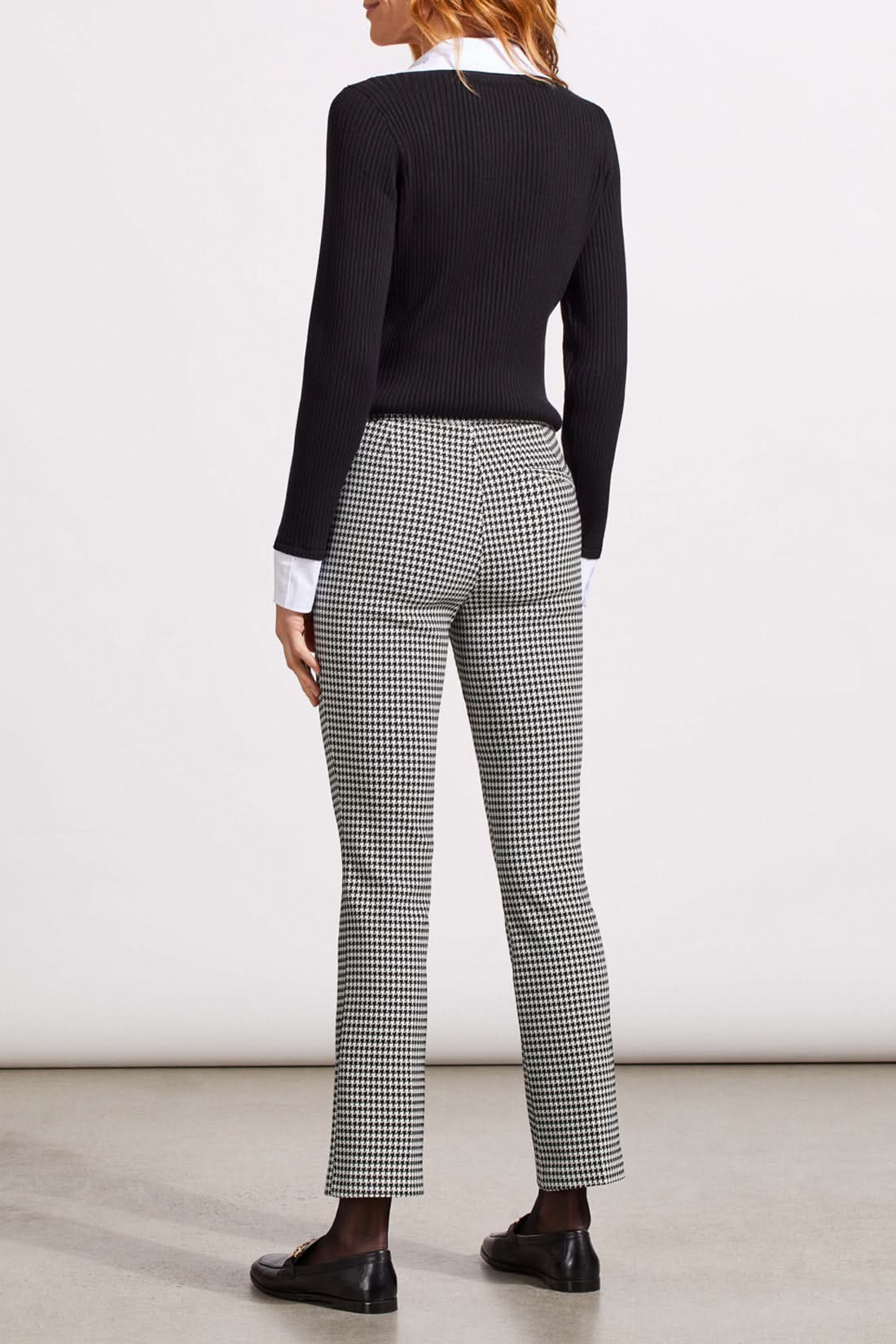 Close-up of a person showcasing the Tribal Houndstooth Pull on Pant, blending comfort and style with its classic houndstooth pattern. They're paired with a black sweater over a white cuffed shirt, revealing part of the torso and one hand.