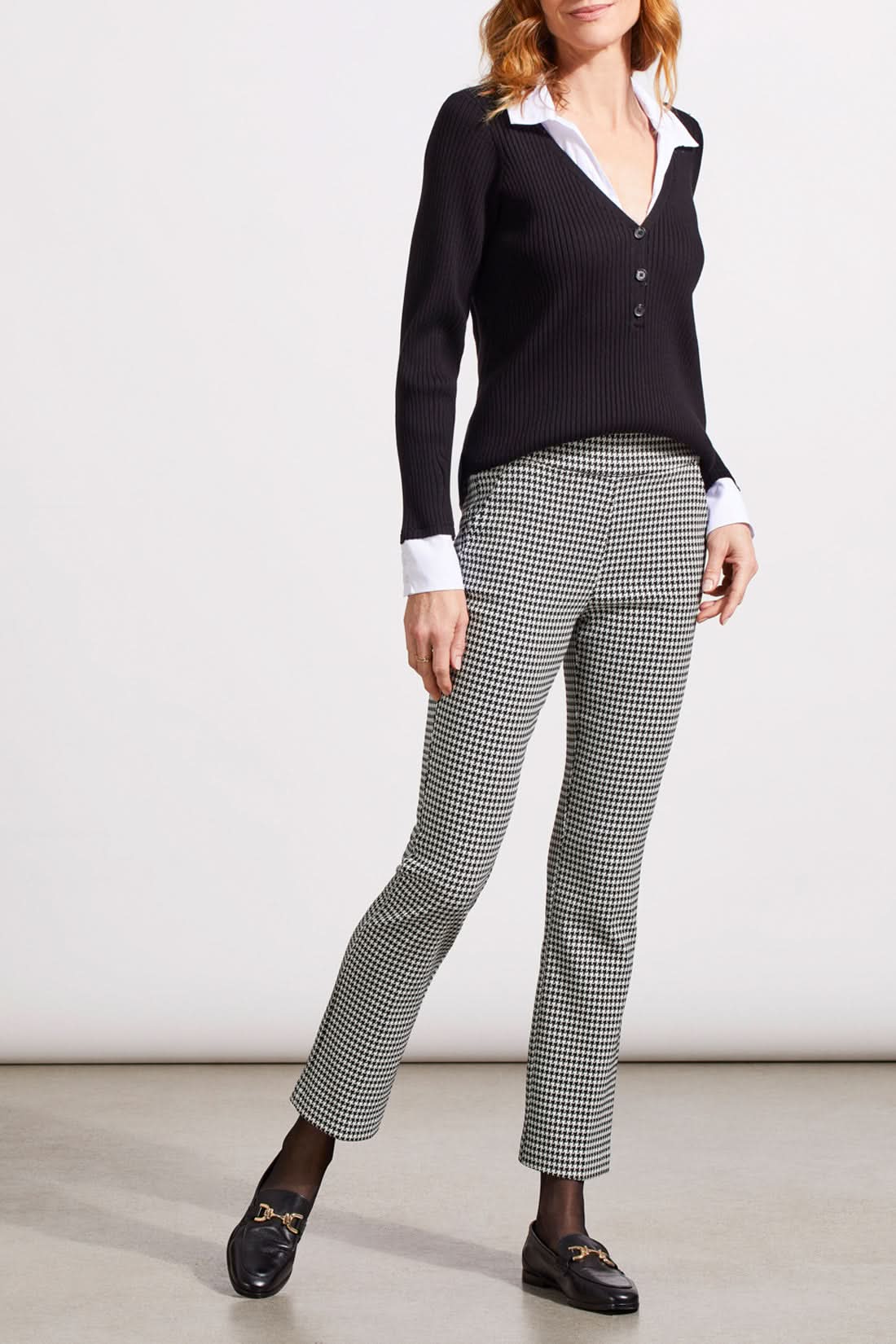 Close-up of a person showcasing the Tribal Houndstooth Pull on Pant, blending comfort and style with its classic houndstooth pattern. They're paired with a black sweater over a white cuffed shirt, revealing part of the torso and one hand.