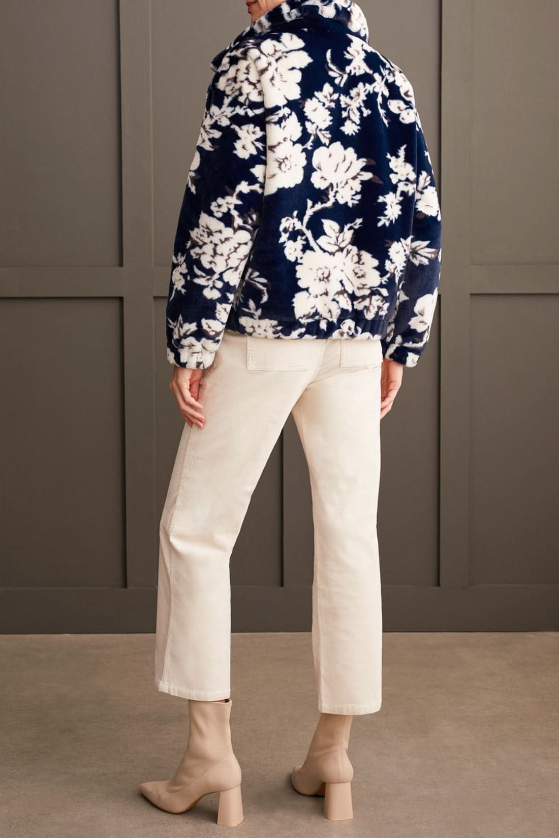 A person wearing the Tribal Zip up Bomber with Lining featuring a floral pattern, along with a dark turtleneck, light-colored pants, and beige boots, stands against a paneled wall.