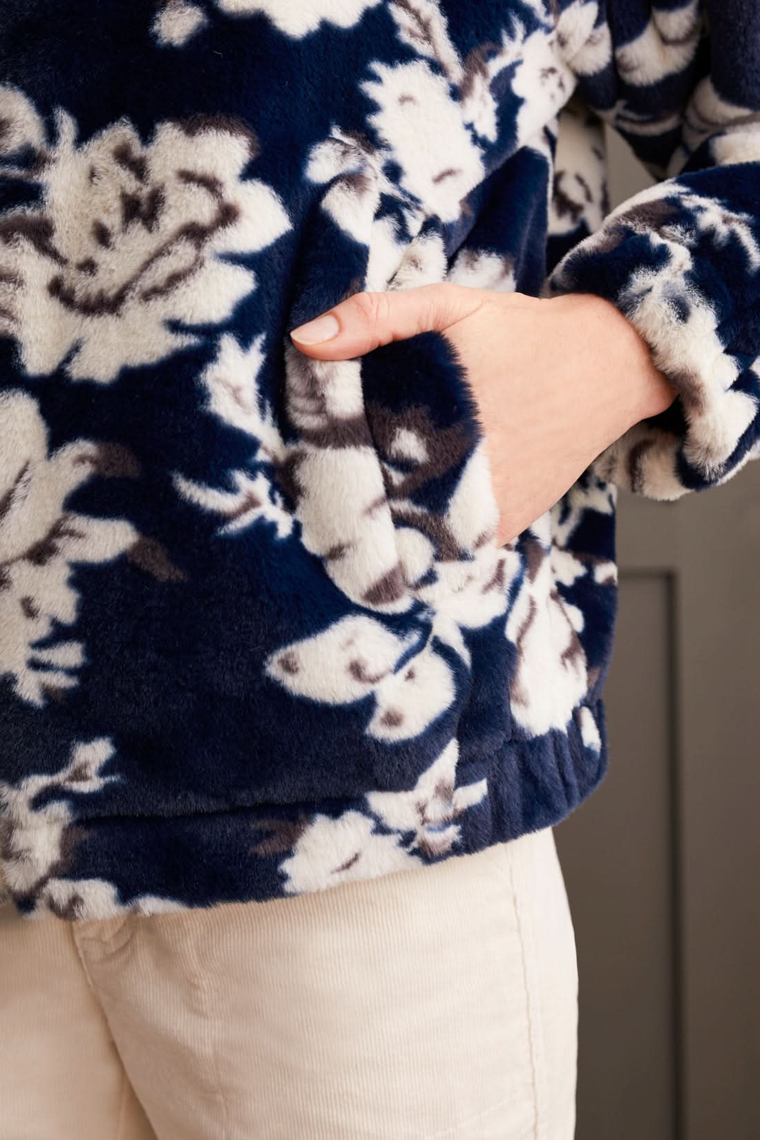 A person wearing the Tribal Zip up Bomber with Lining featuring a floral pattern, along with a dark turtleneck, light-colored pants, and beige boots, stands against a paneled wall.