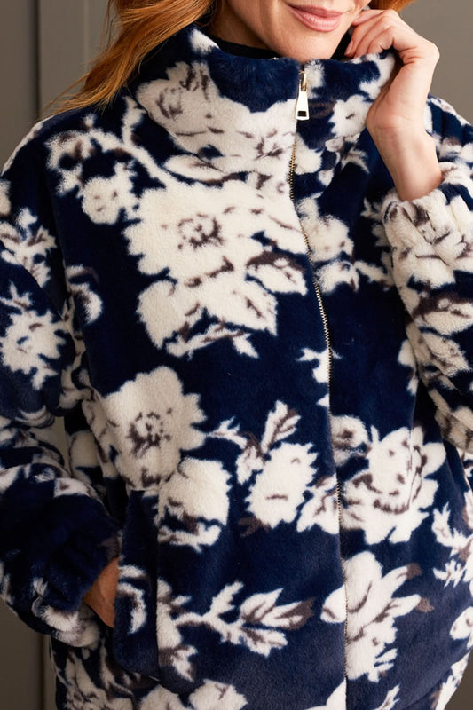 A person wearing the Tribal Zip up Bomber with Lining featuring a floral pattern, along with a dark turtleneck, light-colored pants, and beige boots, stands against a paneled wall.