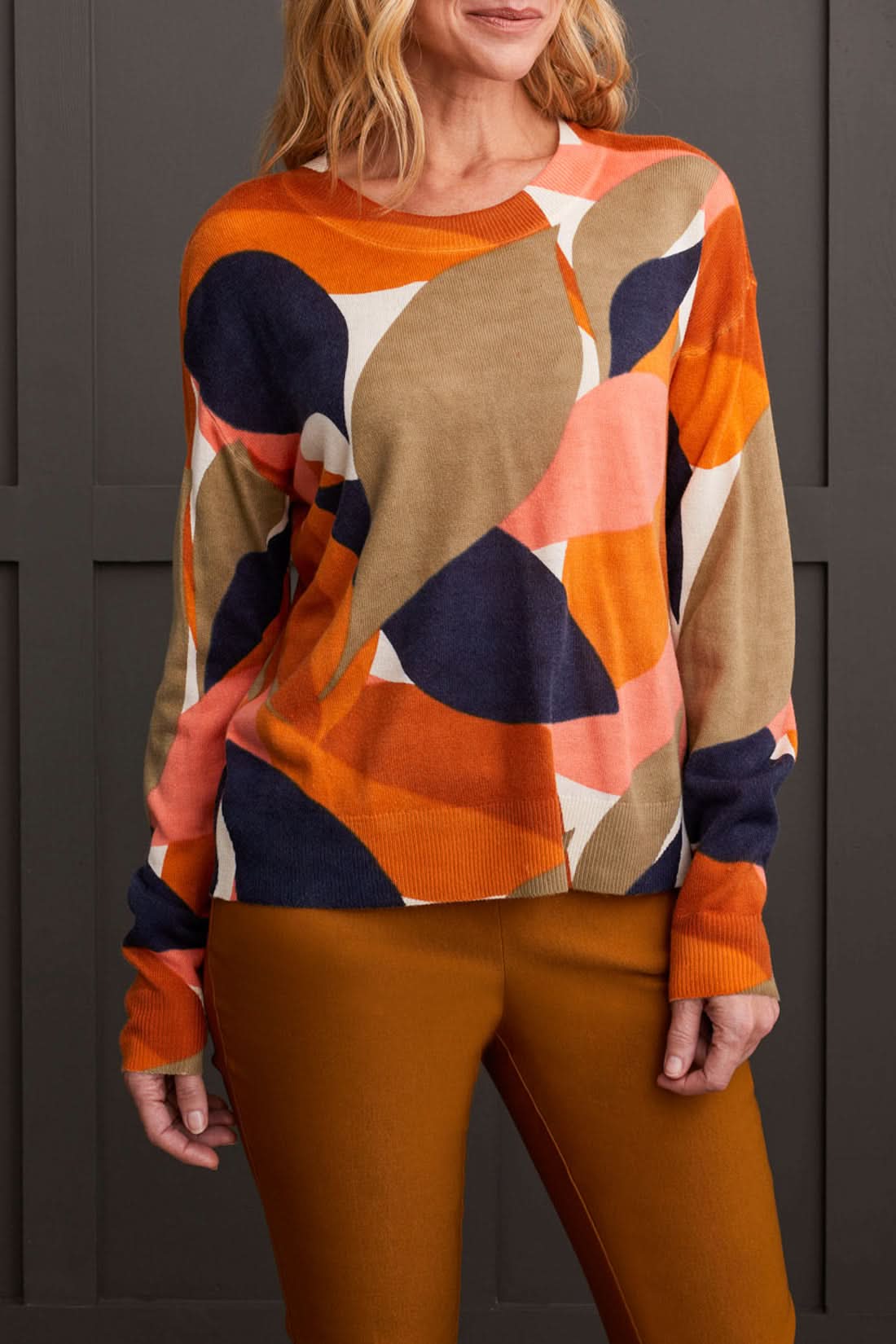 A person wearing a Tribal Long Sleeve Crew Neck Printed Sweater paired with brown pants stands against a dark paneled background.