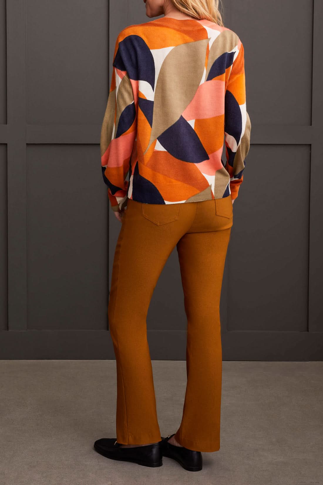 A person wearing a Tribal Long Sleeve Crew Neck Printed Sweater paired with brown pants stands against a dark paneled background.