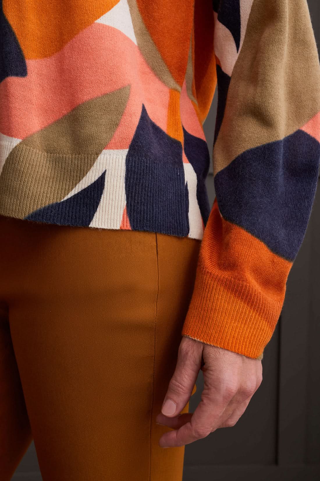 A person wearing a Tribal Long Sleeve Crew Neck Printed Sweater paired with brown pants stands against a dark paneled background.