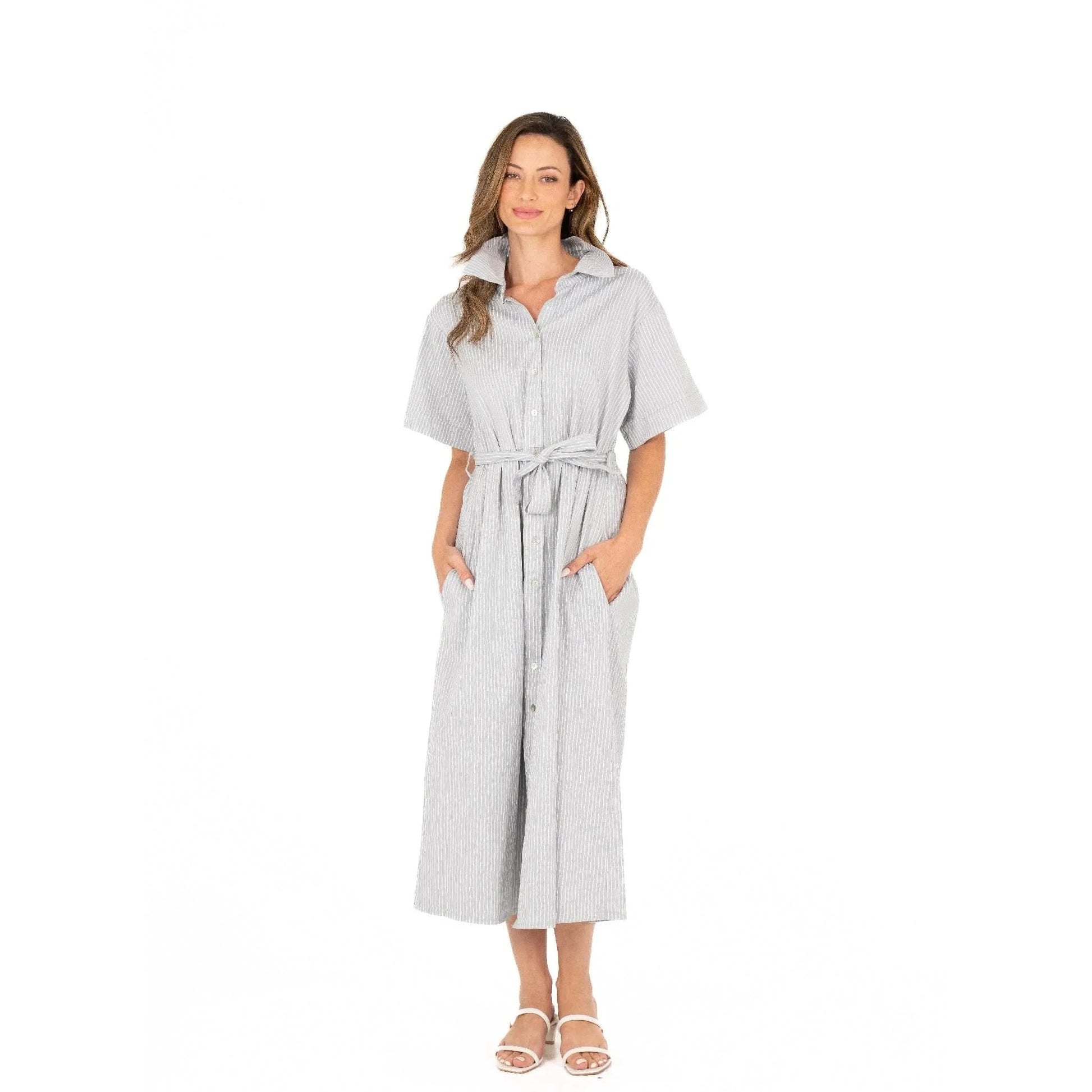 Against a white background, a woman wears the Button Up Short Sleeve Dress with Front Tie by M Made In Italy. Paired with white sandals, her stylish and practical striped dress exudes effortless charm and modern elegance.