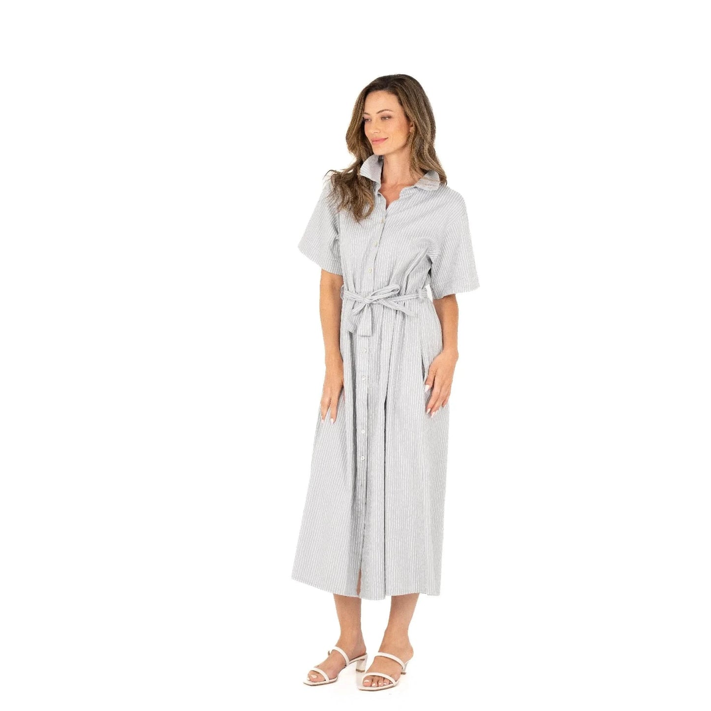 Against a white background, a woman wears the Button Up Short Sleeve Dress with Front Tie by M Made In Italy. Paired with white sandals, her stylish and practical striped dress exudes effortless charm and modern elegance.