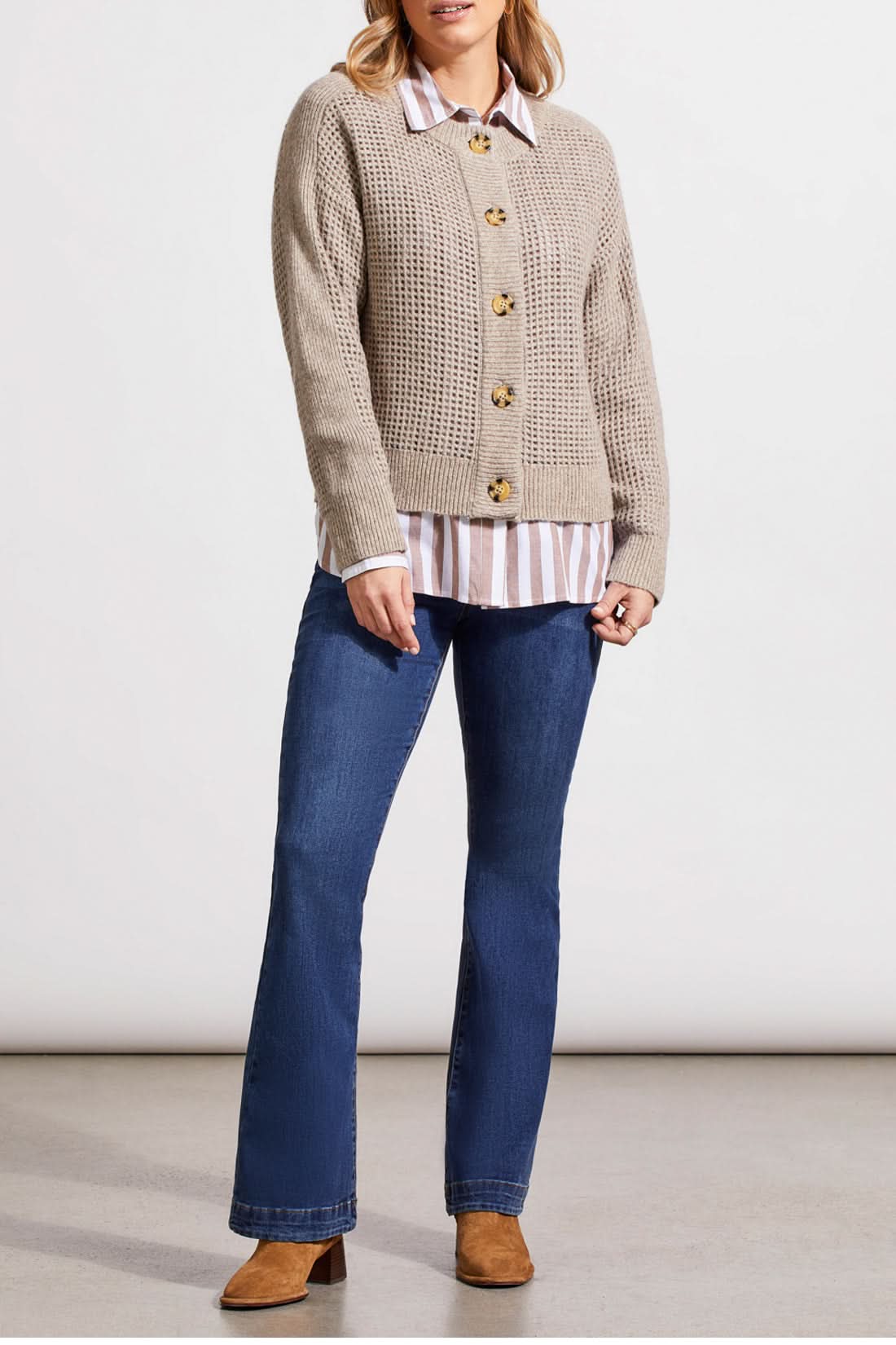 A person wearing a Crew Neck Sweater Cardigan by Tribal in beige, paired with a striped shirt, blue jeans, and brown boots stands against a plain background.