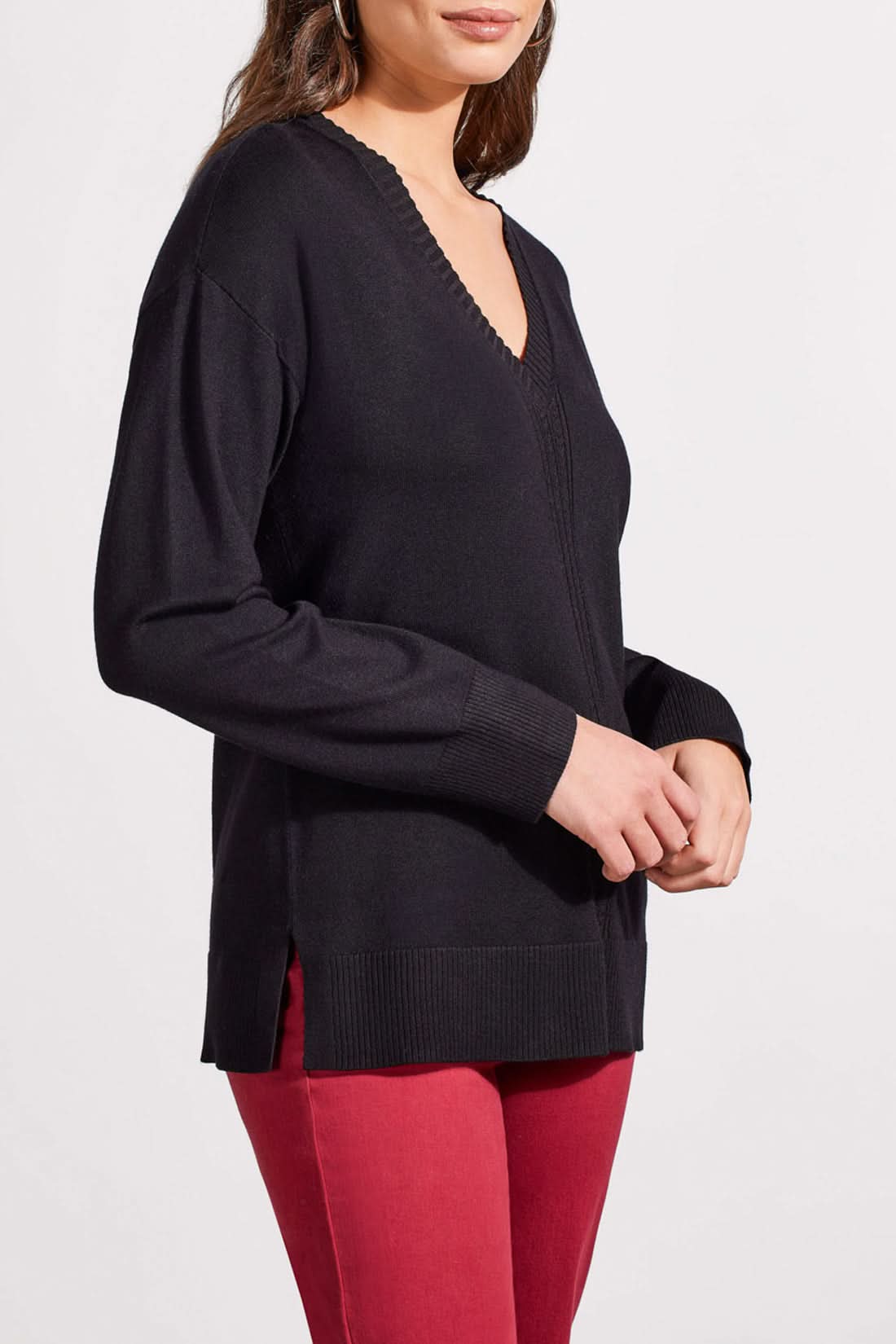 A woman is wearing the Tribal V Neck Sweater with Cables in black and red pants, standing against a plain background. Her hands are clasped in front of her, perfectly capturing the cozy vibe of the fall season.