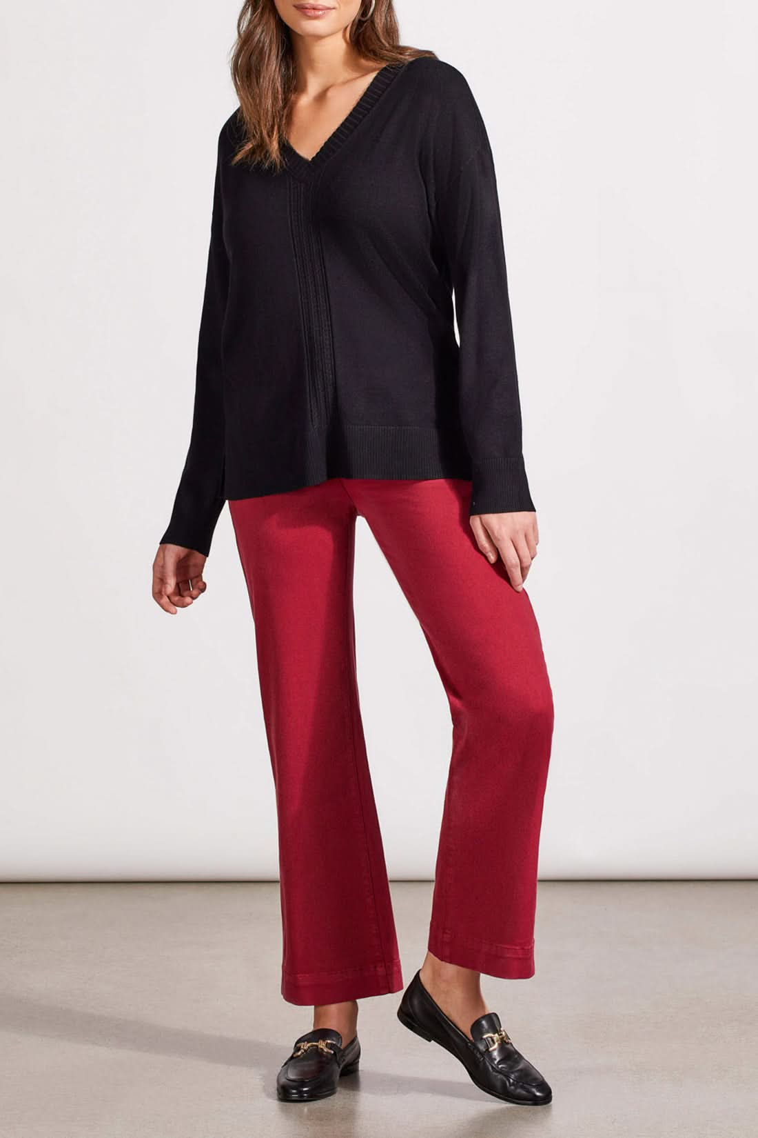 A woman is wearing the Tribal V Neck Sweater with Cables in black and red pants, standing against a plain background. Her hands are clasped in front of her, perfectly capturing the cozy vibe of the fall season.