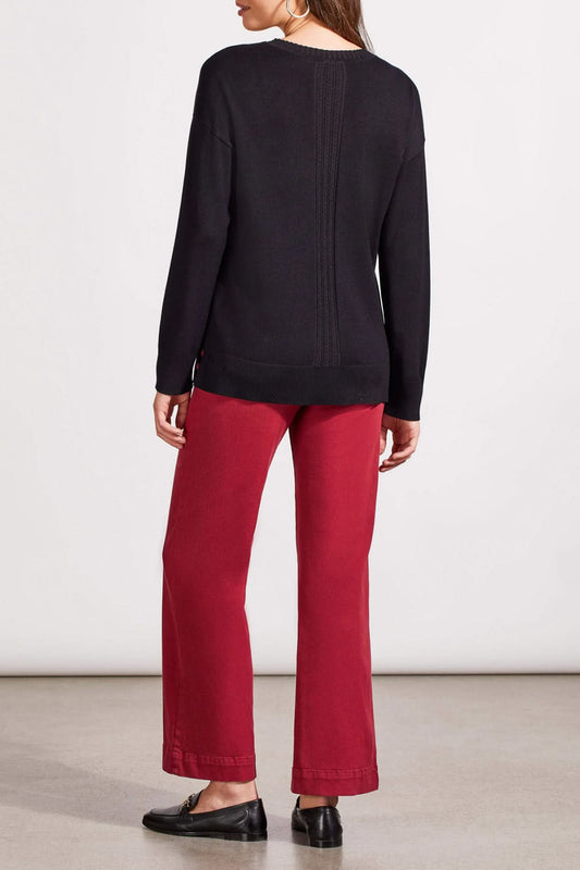 A woman is wearing the Tribal V Neck Sweater with Cables in black and red pants, standing against a plain background. Her hands are clasped in front of her, perfectly capturing the cozy vibe of the fall season.