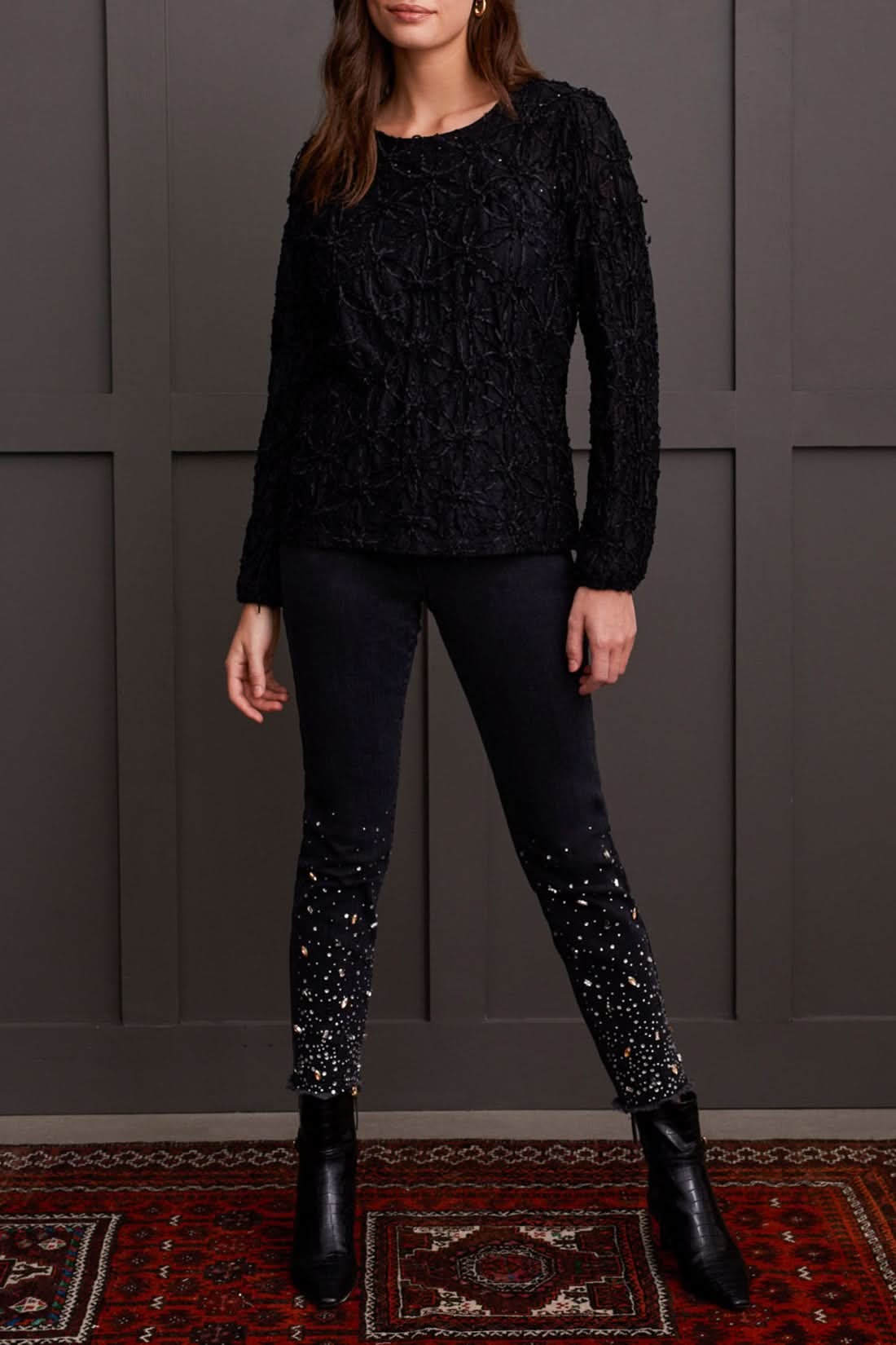 A person wearing a chic Crewneck Top with Integrated Cami by Tribal and dark pants stands gracefully in front of a gray paneled wall.