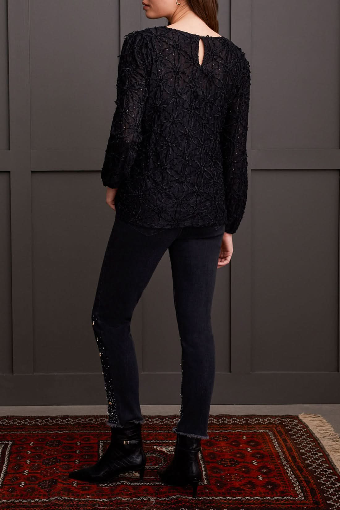 A person wearing a chic Crewneck Top with Integrated Cami by Tribal and dark pants stands gracefully in front of a gray paneled wall.