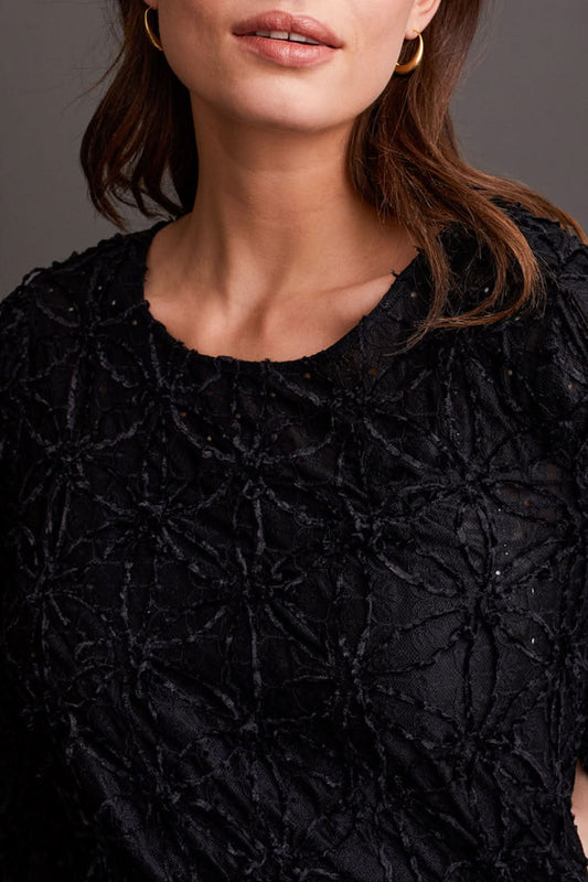 A person wearing a chic Crewneck Top with Integrated Cami by Tribal and dark pants stands gracefully in front of a gray paneled wall.