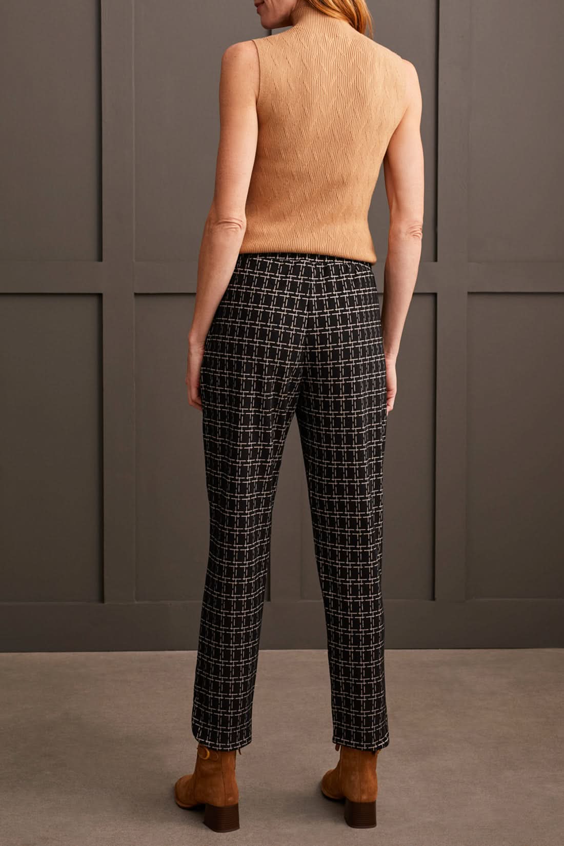 A person stands against a paneled wall, showcasing the Tribal Pull on Ankle Pant with Rounded Hem in black and white checkered print, paired with a sleeveless beige top and brown ankle boots. Perfect for everyday wear.