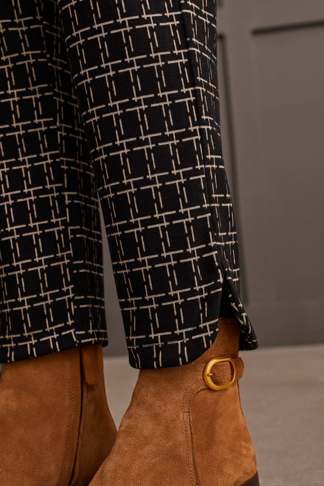 A person stands against a paneled wall, showcasing the Tribal Pull on Ankle Pant with Rounded Hem in black and white checkered print, paired with a sleeveless beige top and brown ankle boots. Perfect for everyday wear.