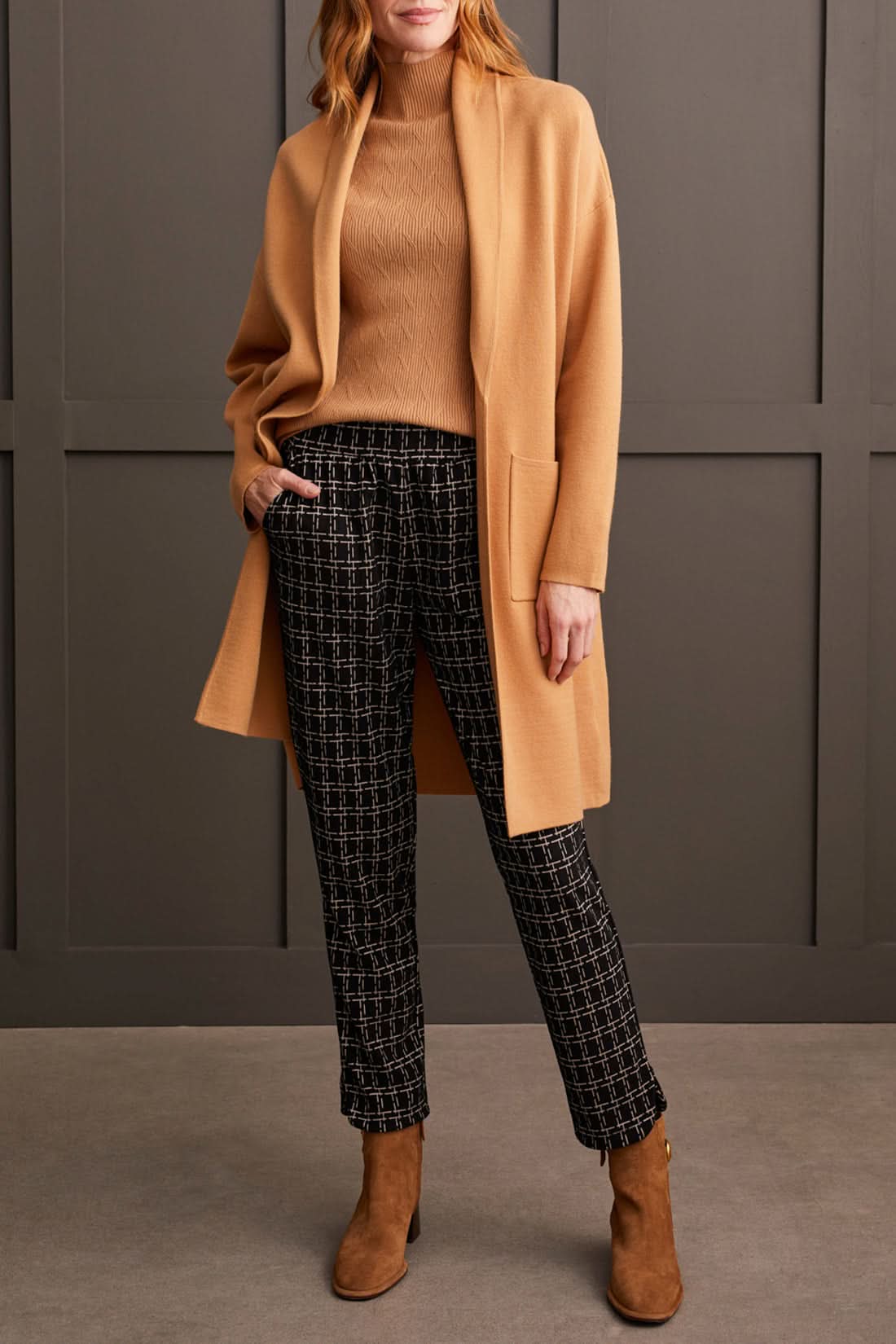 A person stands against a paneled wall, showcasing the Tribal Pull on Ankle Pant with Rounded Hem in black and white checkered print, paired with a sleeveless beige top and brown ankle boots. Perfect for everyday wear.