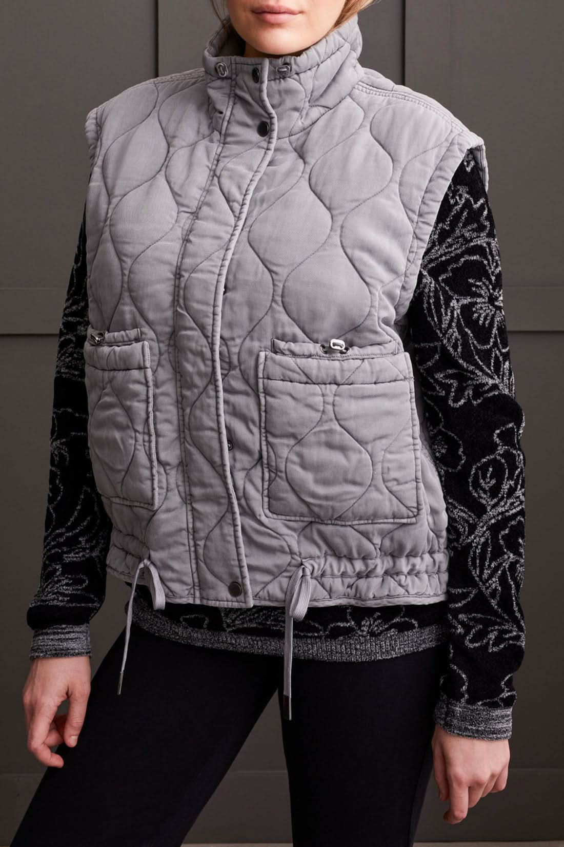 A person is wearing the Tribal Quilted Zip Up Vest in gray, featuring a high collar and two front pockets, over a black long-sleeve shirt with a white floral pattern for extra warmth.
