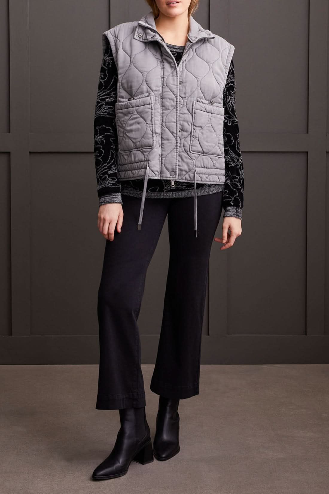 A person is wearing the Tribal Quilted Zip Up Vest in gray, featuring a high collar and two front pockets, over a black long-sleeve shirt with a white floral pattern for extra warmth.