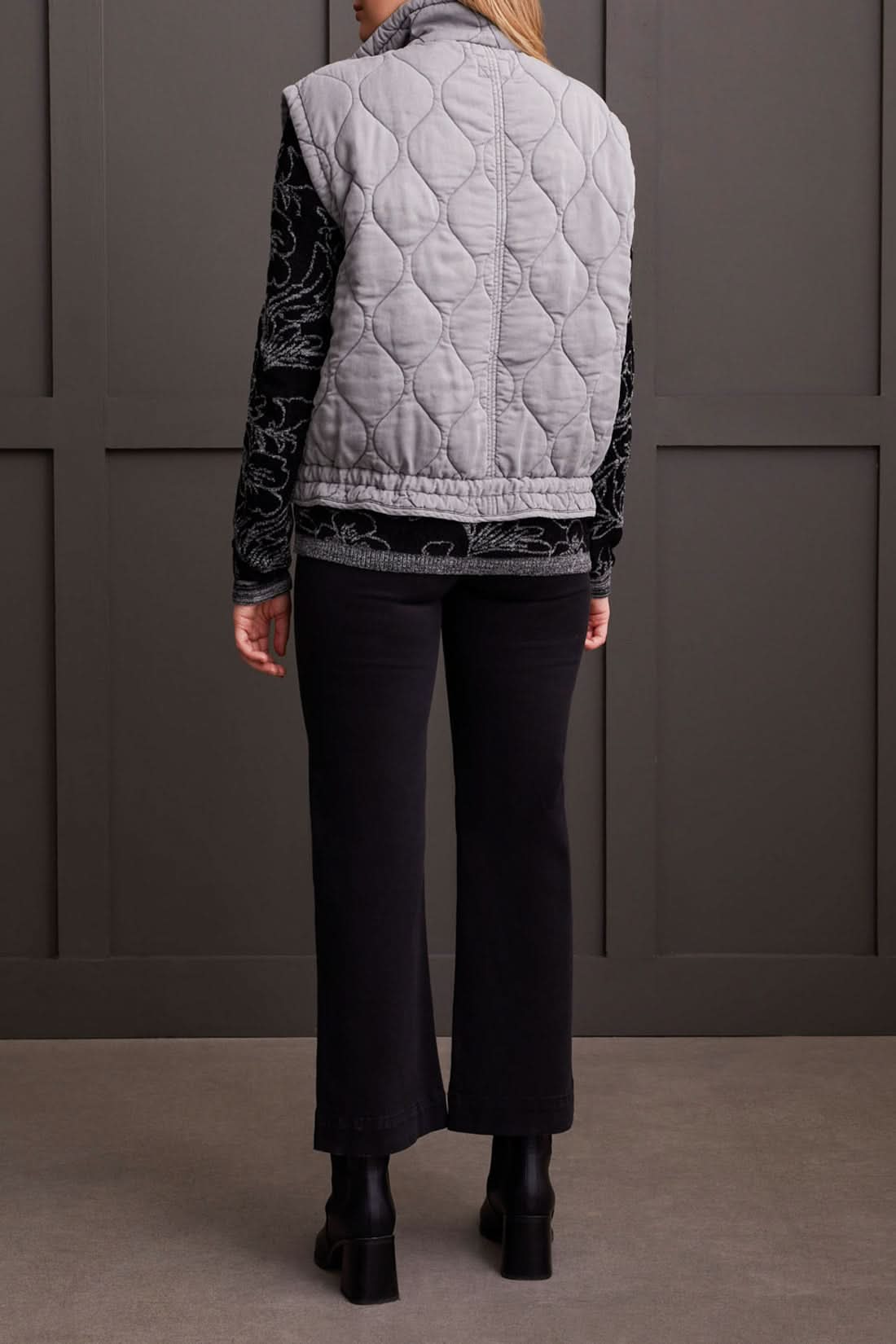 A person is wearing the Tribal Quilted Zip Up Vest in gray, featuring a high collar and two front pockets, over a black long-sleeve shirt with a white floral pattern for extra warmth.