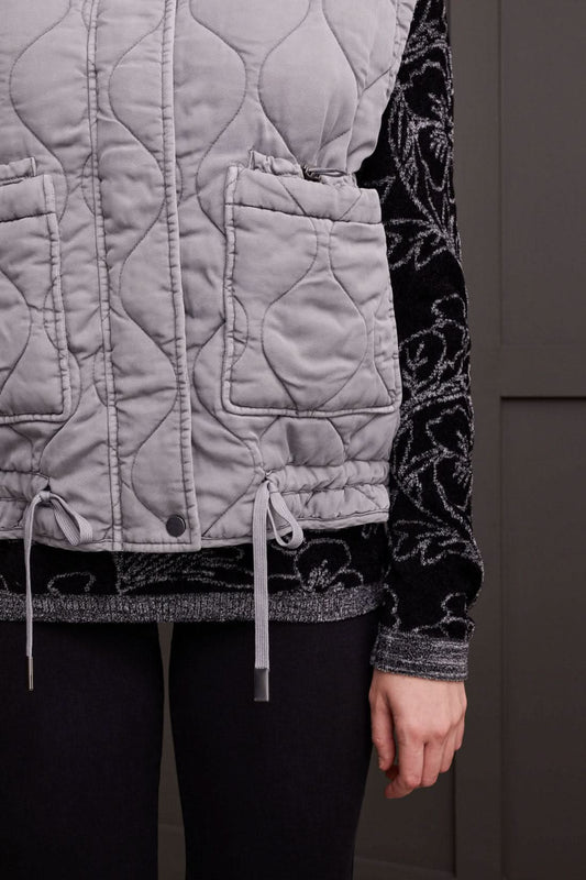 A person is wearing the Tribal Quilted Zip Up Vest in gray, featuring a high collar and two front pockets, over a black long-sleeve shirt with a white floral pattern for extra warmth.