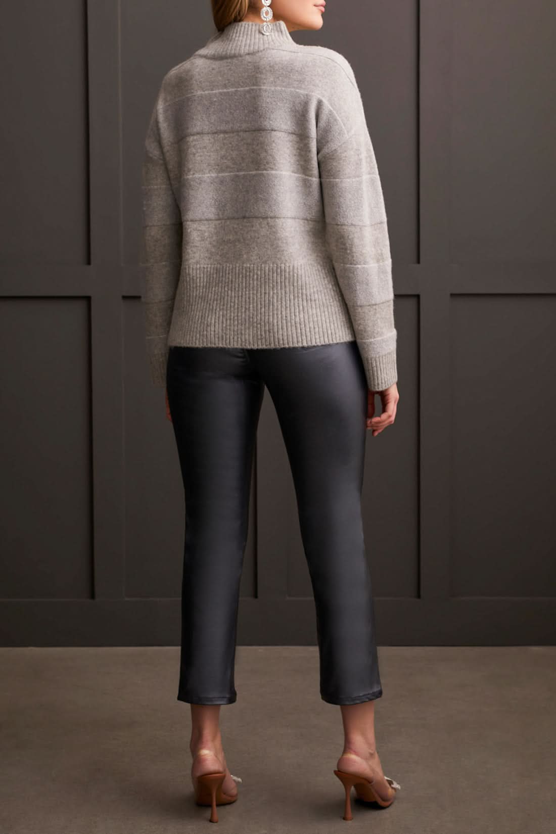 A person is modeling the Tribal Long Sleeve Mock Neck Sweater paired with black leather pants and heeled sandals, creating a chic look against a dark paneled wall—ideal for enhancing your fall wardrobe.