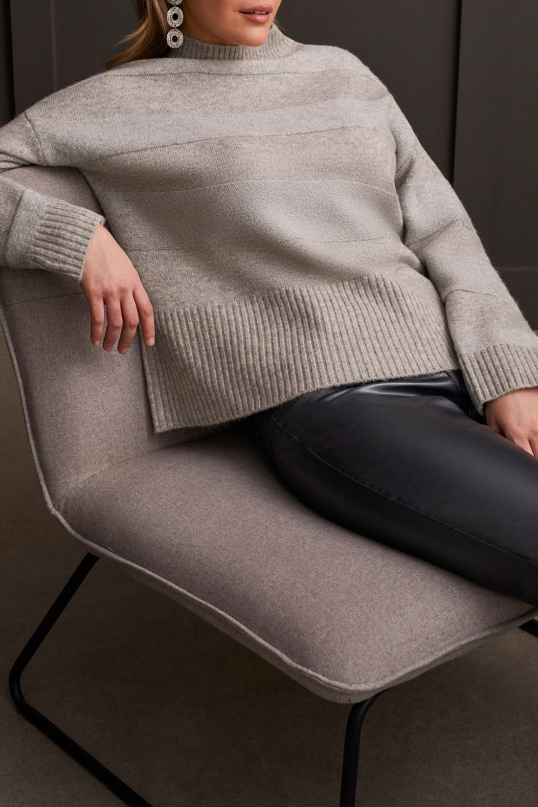 A person is modeling the Tribal Long Sleeve Mock Neck Sweater paired with black leather pants and heeled sandals, creating a chic look against a dark paneled wall—ideal for enhancing your fall wardrobe.